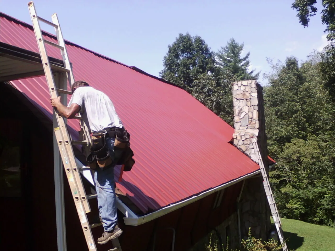 Roofer on a ladder beside a red metal roof and stone chimney — Soffit & Fascia Repair by Roofing Brothers in Buda